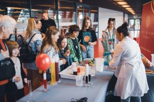 Zdjęcie przedstawia grupę osób oglądających pokaz pokazy suchego lodu i ciekłego azotu, wykonany przez edukatorkę Epi-Centrum Nauki. Pokaz odbył się na stadionie miejskim.